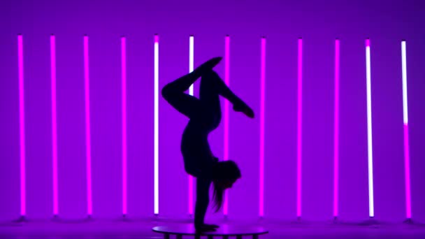 A girl against the backdrop of neon lights. The girl performs a handstand, demonstrates gymnastic movements with her legs, bends and lowers one leg while making a bridge. Dark silhouette. Slow motion. — Stock Video