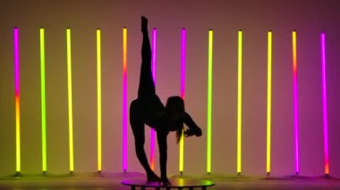 Bir jimnastikçi bacağını kaldırır, ellerine yaslanır, sırtını büker ve bir eliyle stüdyodaki yükseltilmiş bacağa uzanır ve çok renkli neon tüplerin arka planına uzanır. Koyu renk siluet. Yavaş çekim.