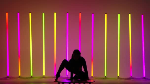Eine junge Frau macht Handstand, macht einen Winkelsprung, bewegt ihre Beine und beugt sie kniend im Studio vor dem Hintergrund bunter Neonlichter. Dunkle Silhouette. Zeitlupe. — Stockvideo