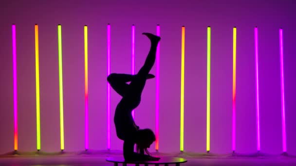 Zijaanzicht van een donker silhouet van een turnster die op haar armen staat, haar benen beweegt, een borststandaard doet en zich in de studio splitst tegen de achtergrond van veelkleurige neonbuizen. Langzame beweging. — Stockvideo