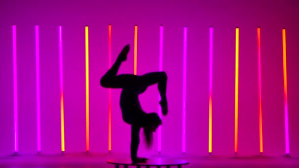 Neon ışıklarının arka planına karşı bir kız. Kız amuda kalkar, bacaklarıyla jimnastik hareketleri gösterir, köprü yaparken bir bacağını eğip indirir. Koyu renk siluet. Yavaş çekim. — Stok video