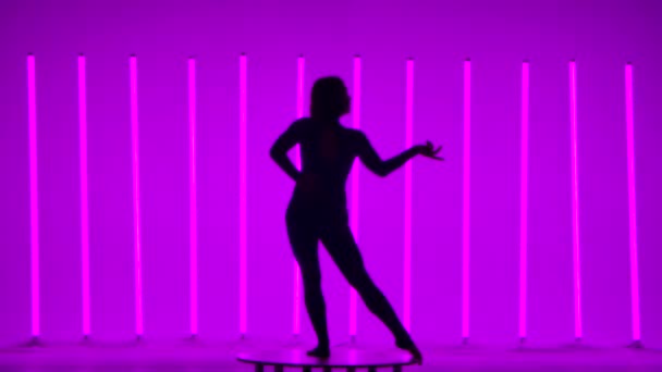 Een slanke atletische vrouw beweegt zich sierlijk tegen de achtergrond van felgekleurde neonbuizen. Het meisje tilt haar been op en doet een verticale split. Donker silhouet. Langzame beweging. — Stockvideo