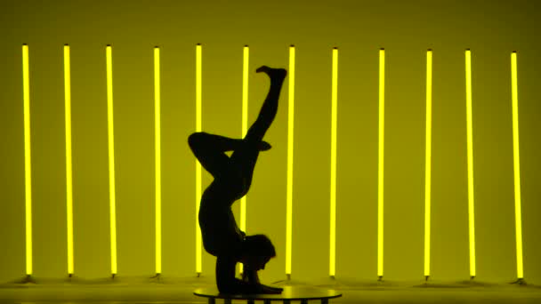 Side view of a dark silhouette of a gymnast who stands on her arms, moves her legs, does a cheststand and splits in the studio against the background of multicolored neon tubes. Slow motion. — Stock Video