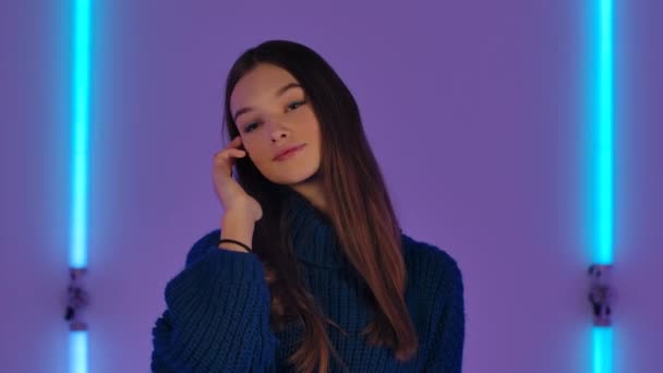 Portrait of fashionable model posing in dark studio against backdrop of bright neon lights. Pretty young woman in blue sweater. Futuristic fashion advertisement. Close up. Slow motion. — Stock Video