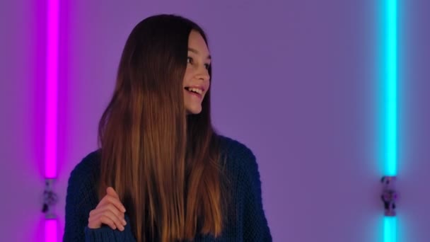 Retrato de modelo de moda bailando alegremente. Mujer morena joven con el pelo largo en suéter azul posa contra el fondo de luces de neón brillantes en el estudio oscuro. De cerca. Movimiento lento. — Vídeos de Stock