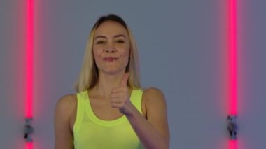 Moda modelinin portresi gülümseyen, baş parmak işareti yapan ve tabela yapan parlak neon ışıkların arka planında uzun saçlı genç sarışın bir kadın. Kapatın. Yavaş çekim.