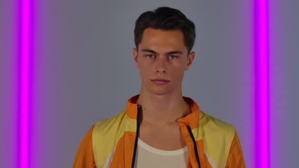 Portrait of stylish guy looking directly at the camera. Male fashion model in yellow jacket poses in dark studio against the backdrop of bright neon lights. Close up. Slow motion. — Stock Video