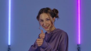 Gülümseyen ve iyi niyet gösteren genç çekici bir kadının portresi. Mor kapüşonlu manken, karanlık stüdyoda parlak neon ışıkların arka planında poz veriyor. Kapatın. Yavaş çekim.