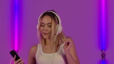 Moda modelinin portresi, büyük beyaz kulaklıklarla dans etmek ve müzik dinlemekten zevk almaktır. Genç sarışın kadın parlak neon ışıkların arkasında poz veriyor. Kapatın. Yavaş çekim.