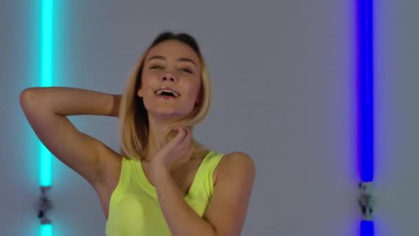 Ritratto di modella alla moda che balla e posa civettuola per la macchina fotografica. Giovane donna bionda in giallo casual top posa sullo sfondo di luminose luci al neon in studio scuro. Chiudete. Rallentatore. — Video Stock