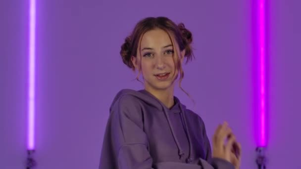 Porträt einer jungen attraktiven Frau, die vor der Kamera posiert. Modemodel im lila Kapuzenpulli vor hellem Neonlicht im dunklen Studio. Aus nächster Nähe. Zeitlupe. — Stockvideo