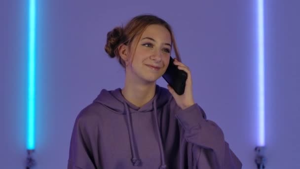Портрет молодої привабливої жінки, що розмовляє для мобільного телефону і радіє. Модна модель в фіолетовому светрі позує на тлі яскравих неонових вогнів у темній студії. Близько. Повільний рух . — стокове відео