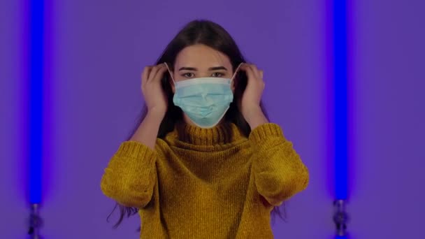 Retrato de mujer joven linda se pone y luego se quita la máscara de protección médica. Modelo de moda con el pelo largo posa contra el fondo de luces de neón brillantes en estudio oscuro. De cerca. Movimiento lento. — Vídeo de stock