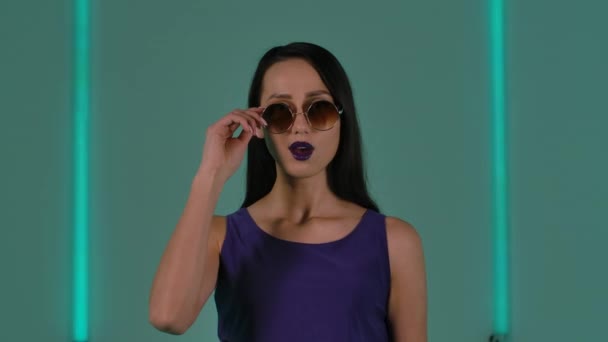 Porträtt av ung snygg kvinna i solglasögon tittar på kamera med wow kvinnliga koketteri. Mode modell med lila läppstift utgör mot bakgrund av ljusa neonljus. Närbild. Långsamma rörelser. — Stockvideo