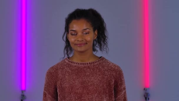 Retrato de una joven afroamericana mirando a la cámara y sonriendo. Modelo de moda en suéter marrón posa sobre fondo de luces de neón brillantes en estudio oscuro. De cerca. Movimiento lento. — Vídeos de Stock