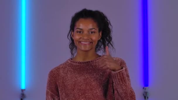 アフリカ系アメリカ人の若い女性の肖像画笑顔と親指をジェスチャーを表示します。ブラウンセーターのファッションモデルは、ダークスタジオの明るいネオンを背景にポーズをとります。閉めろ。スローモーション. — ストック動画