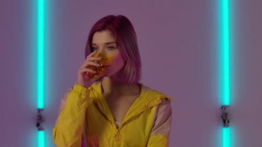 Genç ve şık bir kadının portresi camdan su içiyor sonra da şaşırmış ve şok olmuş bir ifadeyle tükürüyor. Moda modeli parlak neon ışıkların arka planında poz veriyor. Kapatın. Yavaş çekim