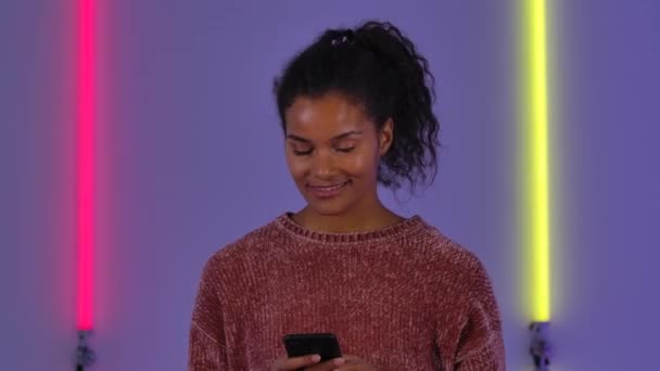 Retrato de afro-americana jovem está enviando mensagens no celular e se alegrar. Modelo de moda em suéter marrom posa contra fundo de luzes de néon brilhantes no estúdio escuro. Fecha. Movimento lento. — Vídeo de Stock