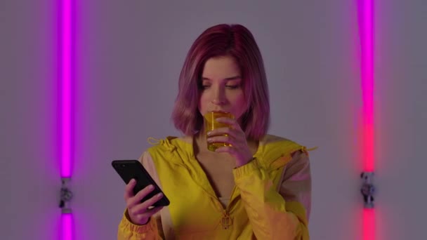 Portrét mladé stylové ženy textuje na telefon a pitnou vodu, vyplivne ji s úžasem a nadšením. Módní model pózuje proti jasným neonovým světlům. Zavřít. Zpomalený pohyb — Stock video