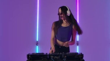 Güneş gözlüklü ve büyük beyaz kulaklıklı genç bir kadının portresi DJ turntable 'da dans ediyor ve müziği karıştırıyor. Moda modeli stüdyodaki parlak neon ışıkların arka planında poz veriyor. Yavaş çekim.