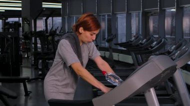 Spor salonundaki koşu bandında koşan yorgun ve terli genç bir kadın. Spor giyimli bayan sporcu kardiyo çalışmasını bitiriyor ve havlularını çıkarıyor. Spor malzemeleri arka planda fitness için. Kapatın. Yavaş çekim.