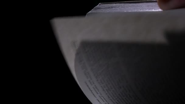 Een man met zijn hand draait de pagina 's van een boek met Engelse tekst in slow motion. Een lichtstraal valt op een boek in een donkere studio op een zwarte achtergrond. Sluiten.. — Stockvideo