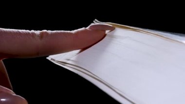 Kız tırnağını kalın bir kitabın sayfalarında gezdiriyor, yavaş çekimde sayfaları çeviriyor. Siyah arka plan üzerine bir kitap. Kapat..