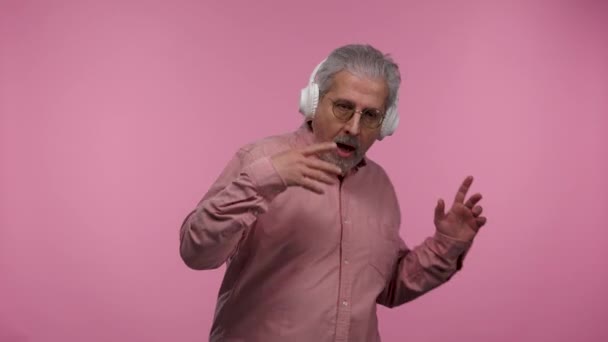 Retrato de un anciano con gafas es divertido bailar y disfrutar de la música en grandes auriculares blancos. Gris pensionista abuelo con barba posando sobre fondo de estudio rosa. — Vídeo de stock