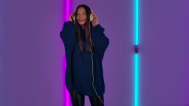 Moda modelinin portresi büyük beyaz kulaklıklarla dans etmek ve müzikten zevk almaktır. Mavi kazaklı uzun saçlı esmer kadın parlak neon ışıkların arkasında poz veriyor. Yavaş çekim.