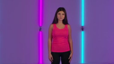 Modaya uygun bir modelin fotoğraf makinesine bakışının portresi. Pembe tişörtlü uzun saçlı esmer kadın karanlık stüdyoda parlak neon ışıkların arka planında poz veriyor. Yavaş çekim.