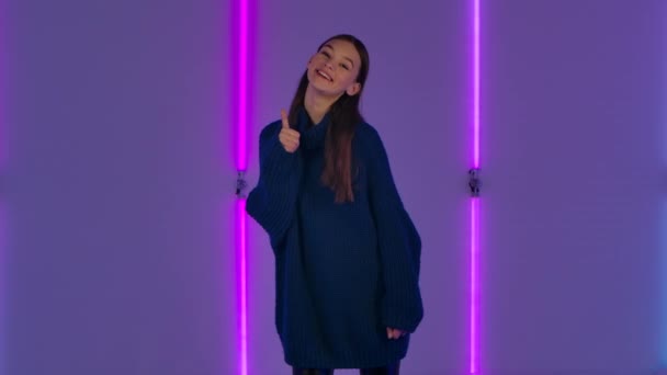 Porträtt av fashionabla modell leende och visar tummen upp gest. Ung brunett kvinna med långt hår i blå tröja utgör mot bakgrund av ljusa neon ljus i mörk studio. Långsamma rörelser. — Stockvideo