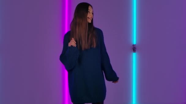 Retrato de modelo de moda bailando alegremente. Mujer morena joven con el pelo largo en suéter azul posa contra el fondo de luces de neón brillantes en el estudio oscuro. Movimiento lento. — Vídeos de Stock