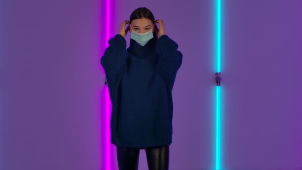 ファッショナブルなモデルの肖像画は、医療保護マスクに置きます。青いセーターの長い髪をした若いブルネットの女性は、暗いスタジオで明るいネオンの背景を背景にポーズをとります。スローモーション. — ストック動画