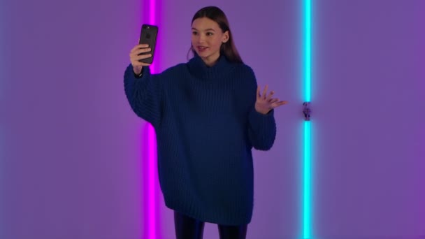 Porträtt av fashionabla modell talar för videochatt med mobiltelefon och glädjas. Ung brunett kvinna med långt hår i blå tröja utgör mot bakgrund av ljusa neonljus. Långsamma rörelser. — Stockvideo