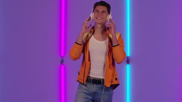 Portrait of stylish guy is dancing and enjoying music in big white headphones. Male fashion model in yellow jacket poses in dark studio against the backdrop of bright neon lights. Slow motion. — Stock Video