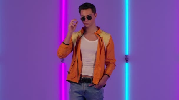 Porträt eines stylischen Typen, der mit Sonnenbrille verspielt in die Kamera blickt. Männermode-Model in gelber Jacke posiert im dunklen Studio vor dem Hintergrund heller Neonlichter. Zeitlupe. — Stockvideo