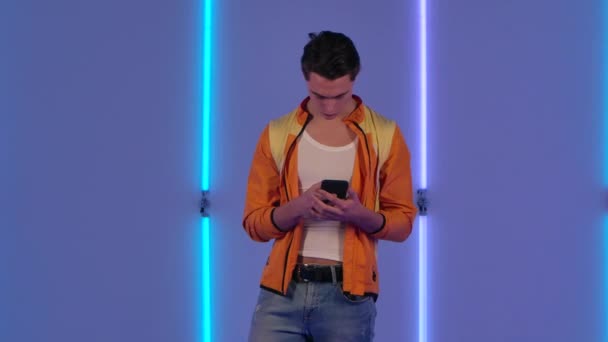 Ritratto di ragazzo alla moda sta messaggiando su cellulare e gioire. Modello di moda maschile in giacca gialla posa in studio scuro sullo sfondo di luminose luci al neon. Rallentatore. — Video Stock