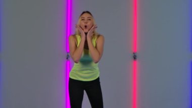 Moda modelinin portresi. Kameraya büyük bir zevkle bakıyor. Sarı bluzlu genç sarışın kadın karanlık stüdyodaki parlak neon ışıkların arka planında poz veriyor. Yavaş çekim.