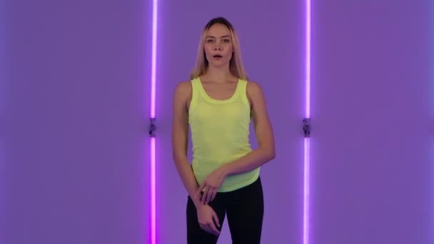 Retrato de modelo de moda mirando a la cámara en serio y luego sonriendo. Mujer rubia joven con el pelo largo en la parte superior casual amarillo posa contra el fondo de luces de neón brillantes. Movimiento lento. — Vídeos de Stock