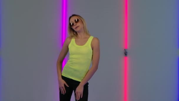 Porträt eines modischen Models mit Sonnenbrille, das kokett für die Kamera posiert. Junge blonde Frau in gelbem, lässigem Top posiert vor hellem Neonlicht im dunklen Studio. Zeitlupe. — Stockvideo