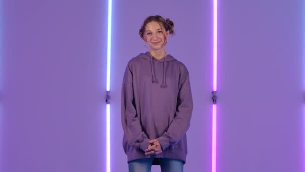 Porträt einer jungen attraktiven Frau, die kokett in die Kamera blickt. Modemodel in lila Kapuzenpulli posiert vor dem Hintergrund heller Neonlichter im dunklen Studio. Zeitlupe. — Stockvideo