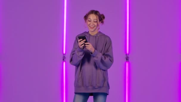 젊고 매력적 인 여자의 모습 이 휴대폰으로 문자 메시지를 보내고 있고 기뻐 합니다. 보라색 후드의 패션 모델은 어두운 스튜디오에 있는 밝은 네온 조명의 배경 과 대조 된다. 느린 동작. — 비디오