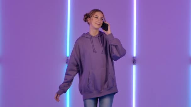 Портрет молодої привабливої жінки, що розмовляє для мобільного телефону і радіє. Модна модель в фіолетовому светрі позує на тлі яскравих неонових вогнів у темній студії. Повільний рух . — стокове відео