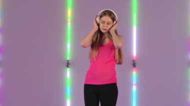 Genç ve güzel bir kadının portresi büyük beyaz kulaklıklarla dans edip müzik dinlemektir. Pembe tişörtlü uzun saçlı manken parlak neon ışıkların arka planında poz veriyor. Yavaş çekim.