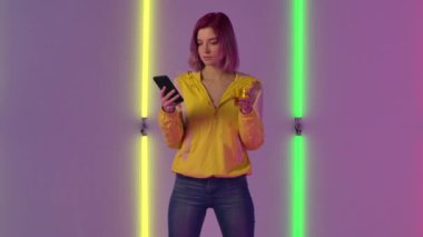 Genç ve şık bir kadının portresi telefonda ve içme suyuyla mesajlaşıyor, şaşkınlık ve coşkuyla tükürüyor. Moda modeli parlak neon ışıklara karşı poz veriyor. Yavaş çekim