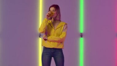 Genç ve şık bir kadının portresi camdan su içiyor sonra da şaşırmış ve şok olmuş bir ifadeyle tükürüyor. Moda modeli parlak neon ışıkların arka planında poz veriyor. Yavaş çekim