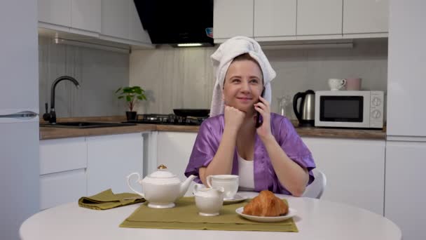 Mujer joven está sentada en la mesa en la cocina, hablando por teléfono, preparándose para el desayuno y beber café. Mujer en bata de baño púrpura y toalla en la cabeza posa sobre el fondo de la sala de luz. — Vídeo de stock