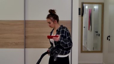 Süpürme yapan, telefona bakan, konuşan ve zaferi kutlayan genç bir kadın. Ev hanımı, ev kıyafetleri içinde, ışık odasına karşı poz veriyor. Kapatın. Yavaş çekim.