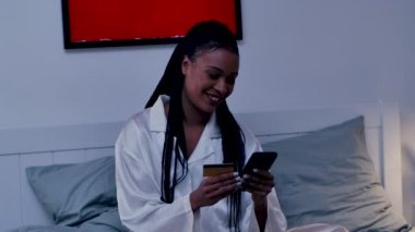 Güzel bir Afrikalı Amerikalı kadın internet üzerinden cep telefonu ve kredi kartı kullanarak alışveriş yapıyor. Siyah kadın pijamalarıyla yatak odasında poz veriyor. Odanın içi aydınlık. Kapatın. Yavaş çekim.