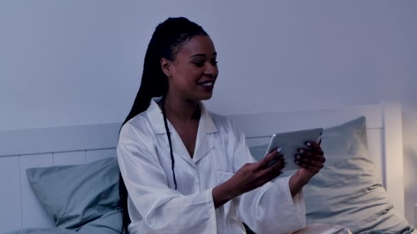 Όμορφη Αφρο-Αμερικανίδα μιλάει σε μια βιντεοκλήση χρησιμοποιώντας ένα ψηφιακό tablet και είναι ευτυχισμένη. Μαύρη γυναίκα που ποζάρει στο κρεβάτι με τις πιτζάμες της. Φωτεινό εσωτερικό του δωματίου στο σπίτι. Κλείσε. Αργή κίνηση. — Αρχείο Βίντεο